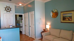 Una sala de estar con un sofá color beige, un banco de madera, una mesita con una lámpara, un reloj de pared y un cuadro enmarcado.