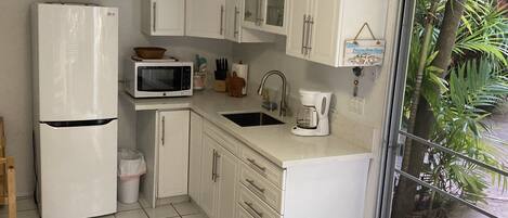 Cocina compacta con armarios blancos, microondas, refrigerador, lavamanos y cafetera.