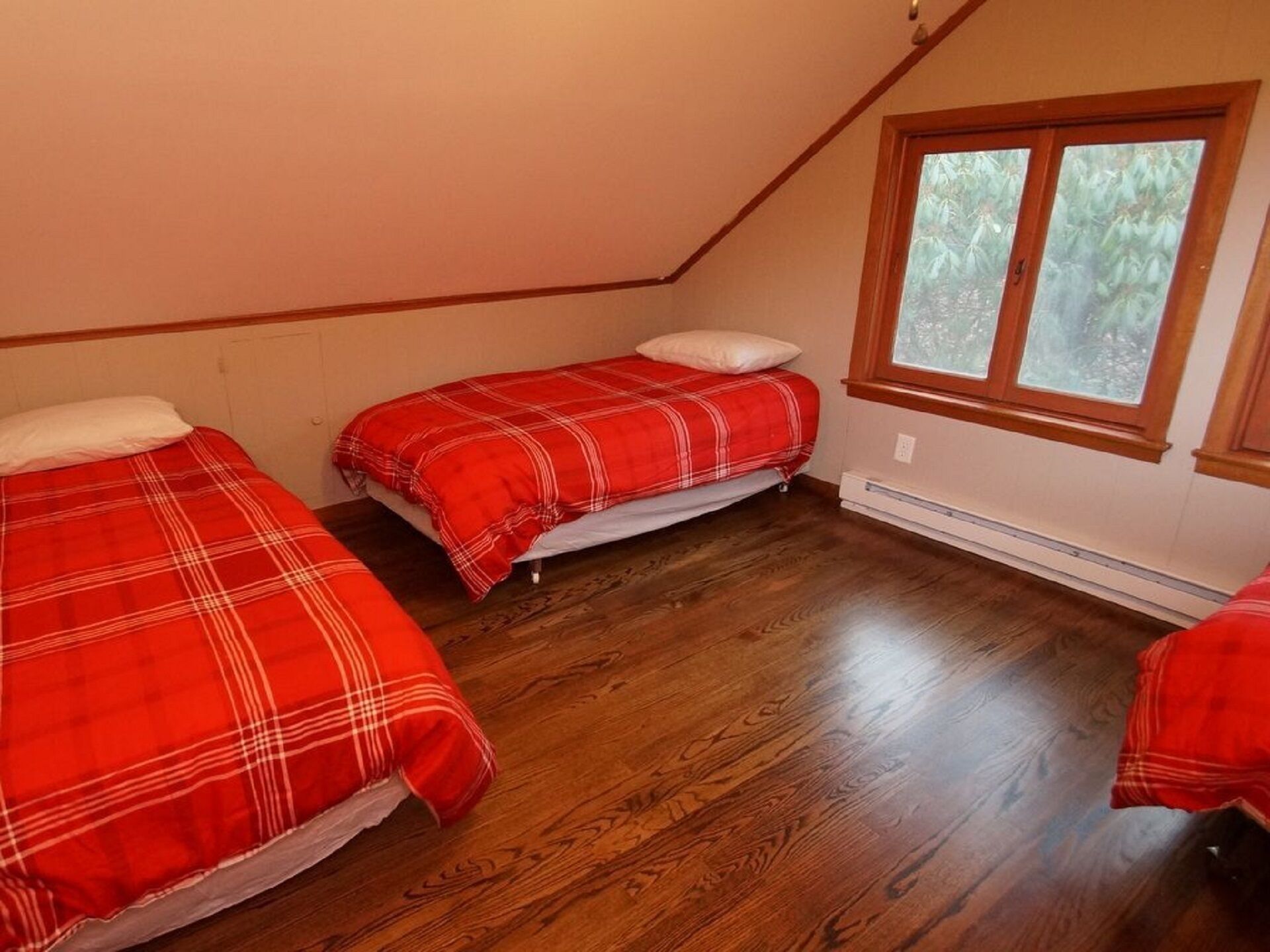 Una habitación con dos camas, suelo de madera, un ventanal con vistas a árboles y techo inclinado.