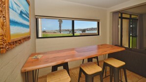 A room with a wooden table and three stools, a large window with a view of a beach and palm tree, and a framed picture on the wall. - Riverview Holiday Waterfront Apt  11 - 2 Storey - Kalbarri, WA (Kalbarri)