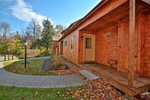 Una cabaña de madera con porche, un sendero de ladrillo y un pequeño jardín con hojas caídas.