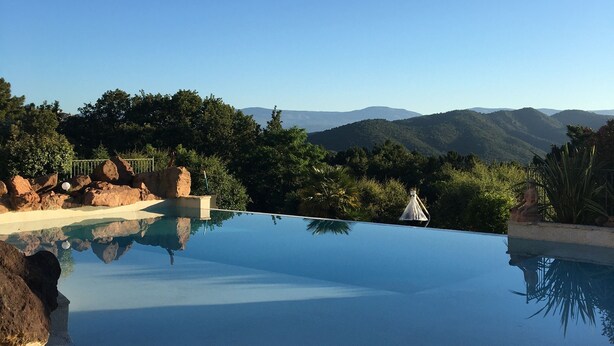 A clear blue swimming pool surrounded by a stone deck, with a view of distant mountains and lush greenery.