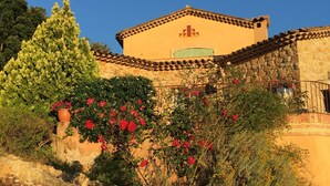 A building with a stone facade and a tiled roof, surrounded by a variety of flowering plants and a large tree.