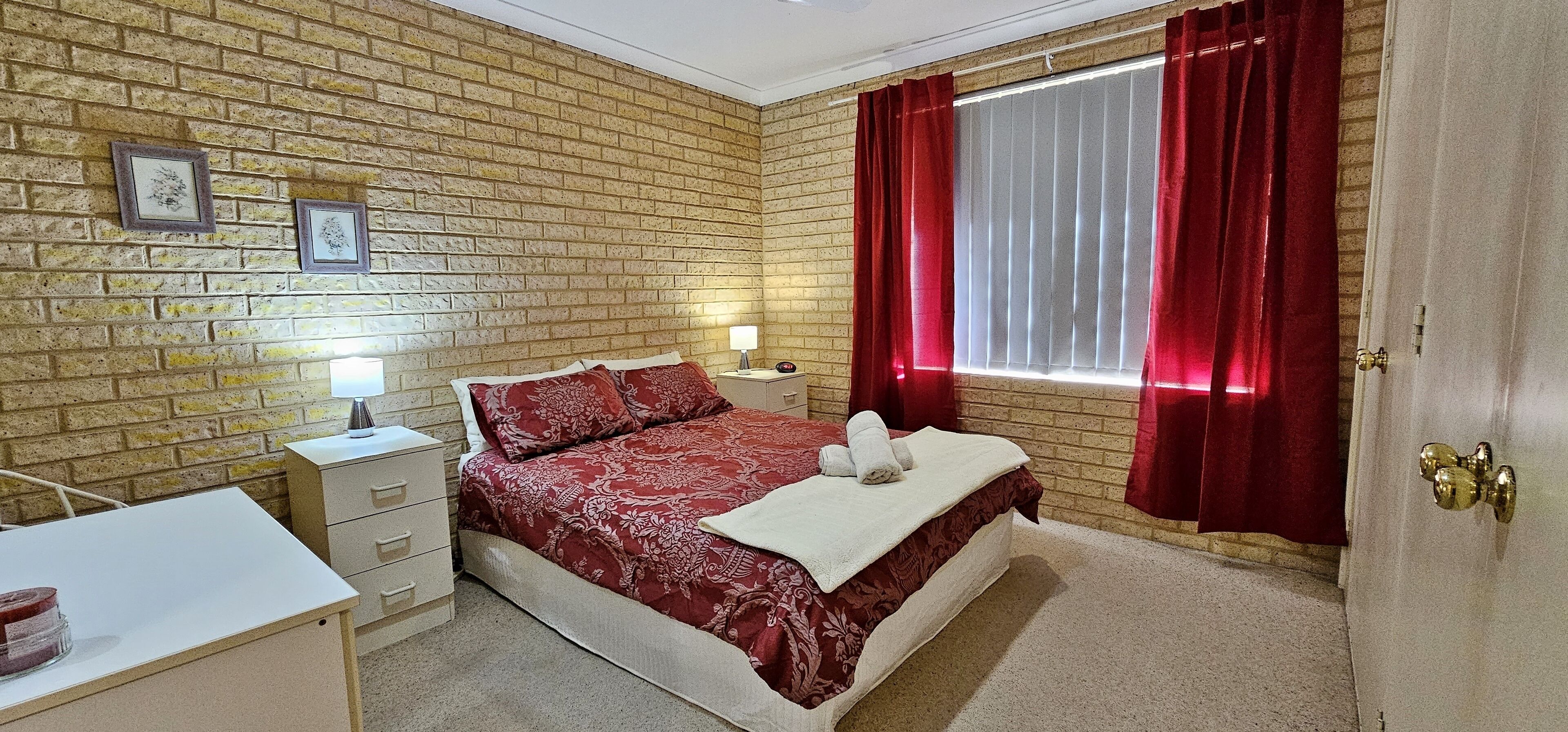A bedroom with a brick-patterned wall, a bed with red bedding, a desk with a lamp, and a window with red curtains.