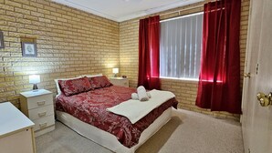Un dormitorio con pared de ladrillos, cama con ropa de cama roja, escritorio con lámpara y ventana con cortinas rojas.