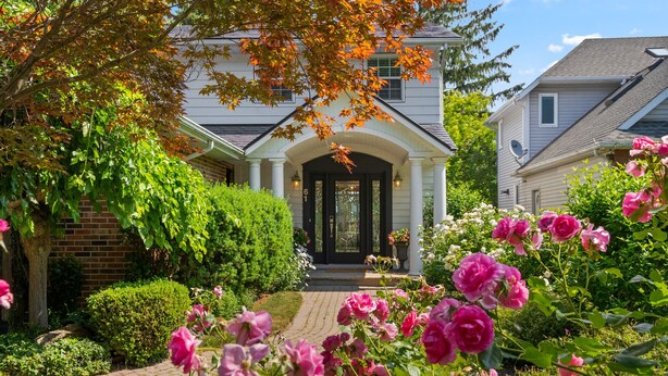 Une maison à deux étages dotée d’un porche abrité, entourée d’un jardin bien entretenu où poussent des rosiers roses.