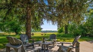 Un espace extérieur aménagé avec des chaises Adirondack et une table ronde, entouré d’arbres et offrant une vue sur un lac.