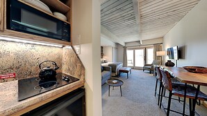 A kitchen with a microwave, kettle, and a sign indicating a sink under the counter. The room has a dining area with a table, chairs, and a TV. - 🌲Timberline 117 Studio Deluxe ~ Hot Tub~Pool~WiFi~Ski-in/out~Local Shuttle🌲 (Snowmass Village)