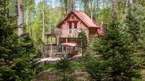 A cabin with a red roof, a balcony, and a stone foundation surrounded by dense forest. - Log Cabin with Private Outdoor Hot Tub (Hot Springs)