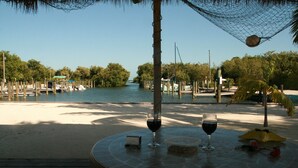 Une table ronde, deux verres à vin et un petit appareil sont disposés au centre, entourés d’une terrasse en bois qui donne sur un plan d’eau où l’on aperçoit des bateaux et des arbres.