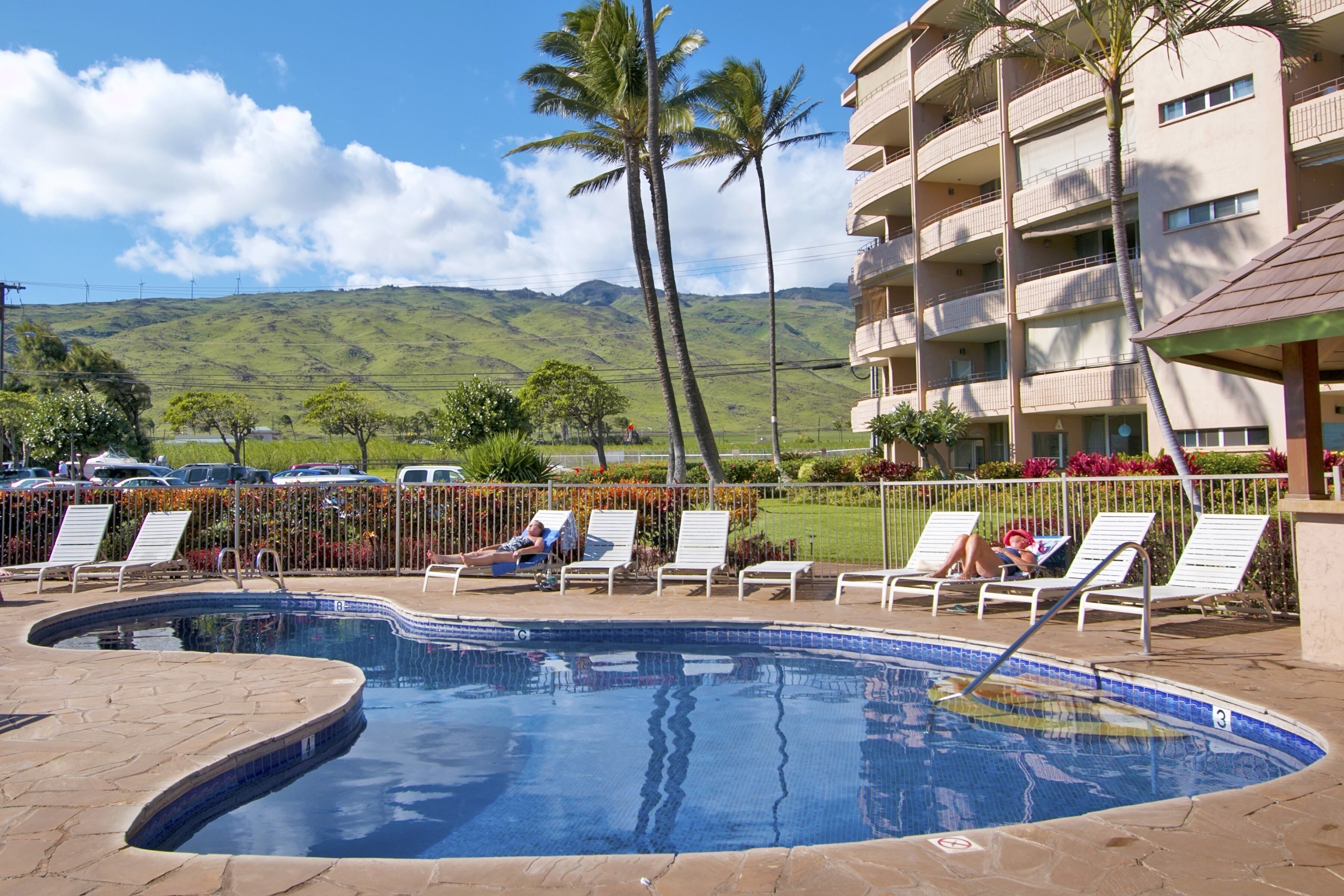 A swimming pool with lounge chairs, a building with balconies, and a view of green hills.