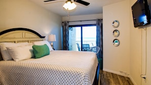 Habitación de hotel con cama, ventilador de techo, televisor montado en la pared y vista al océano a través de la ventana.