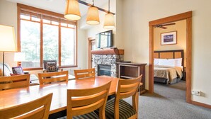 A dining area with a wooden table and chairs, a stone fireplace, and a bed with a headboard in the background.
