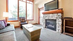 A living room with a stone fireplace, a flat-screen TV mounted above it, a sofa, a coffee table, and a window with blinds.