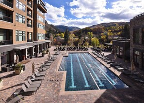 Piscina com espreguiçadeiras e guarda-sóis, rodeada por edifícios e uma paisagem montanhosa.