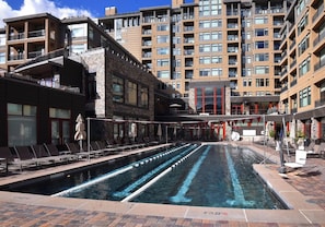 Ein Schwimmbecken mit mehreren Bahnen, umgeben von Liegestühlen und Sonnenschirmen, mit einem mehrstöckigen Gebäude im Hintergrund.
