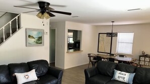 Uma sala de estar com dois sofás de couro preto, um ventilador de teto, uma área de jantar com mesa e cadeiras de madeira, e uma cozinha ao fundo.