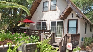 Uma casa rústica com um deck de madeira, um guarda-sol vermelho e diversas plantas.