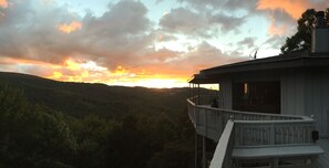 A house with a balcony overlooking a forested valley during sunset.