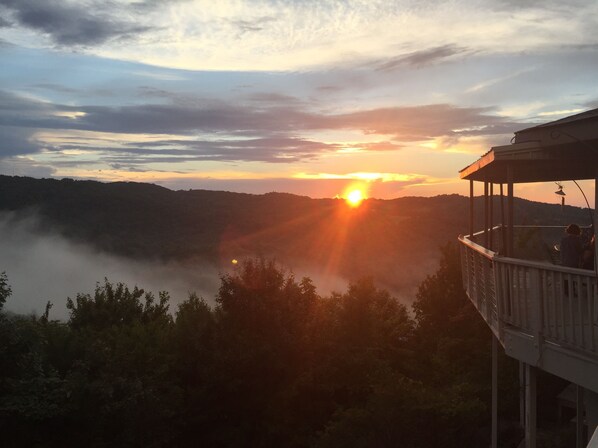 A sunset view from a balcony overlooking a valley with mist and distant hills.