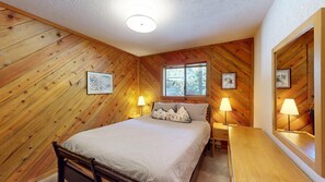 A cozy room with wooden walls, a bed with striped bedding, two bedside tables with lamps, and framed pictures on the walls.