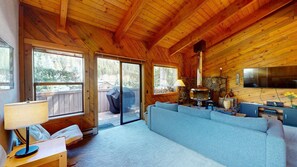 L’intérieur chaleureux d’un chalet avec une cheminée en pierre, un canapé, un téléviseur et une grande fenêtre donnant sur une terrasse.