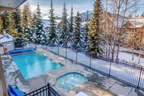 Ein Swimmingpool mit Whirlpool, umgeben von einem Metallzaun und verschneiten Bäumen im Hintergrund.
