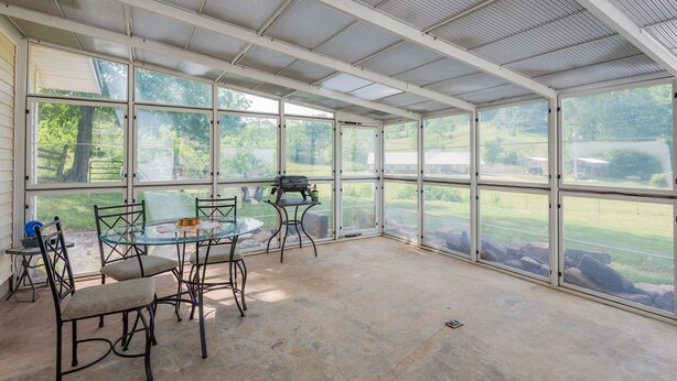 Un patio cubierto con una mesa y sillas de vidrio, ventanas amplias y vistas a un paisaje verde.