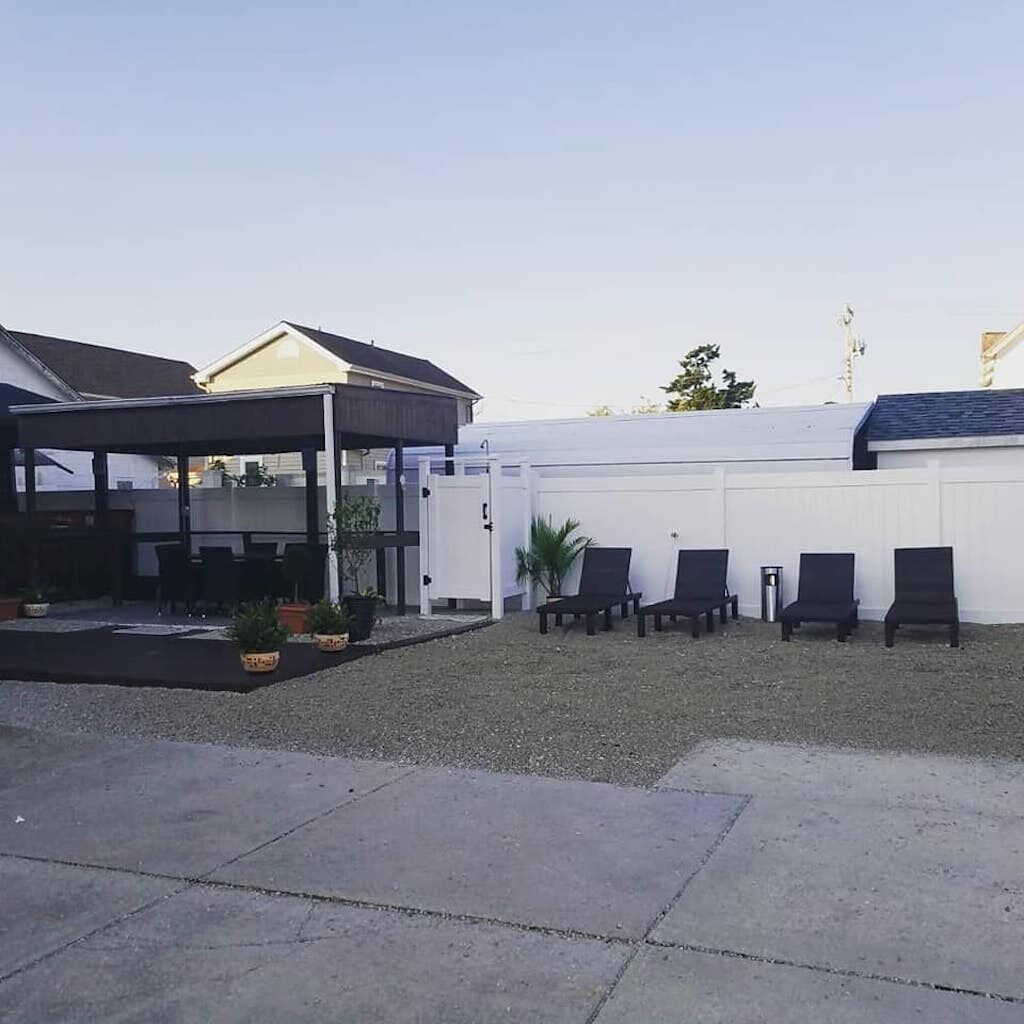 A covered outdoor seating area with chairs and tables, a white fence, and a building with a dark roof.