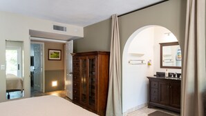 Une chambre avec un grand lit, une armoire en bois, une salle de bain avec un miroir et un lavabo, et une porte donnant sur une autre pièce.