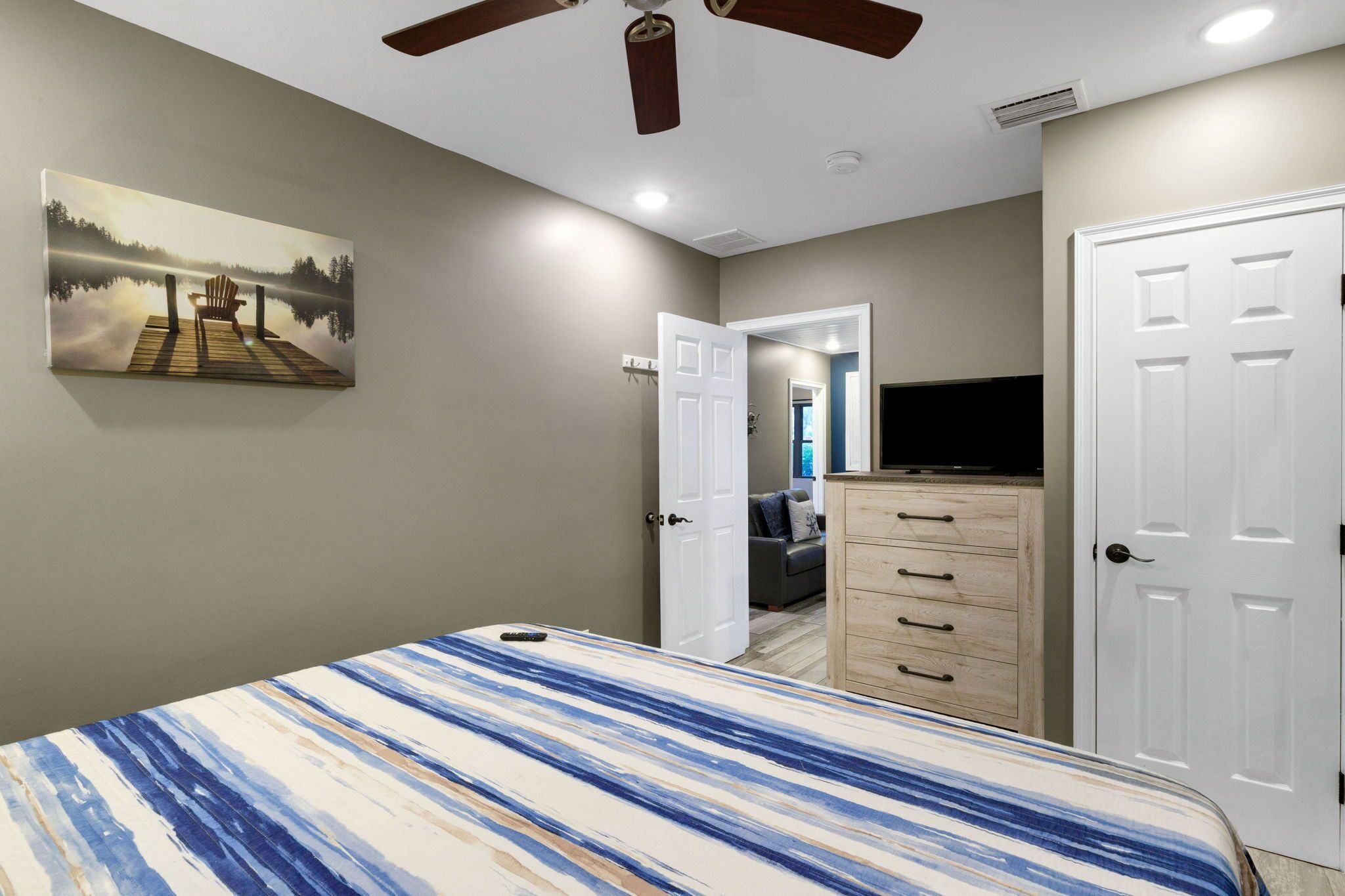 A bedroom with a bed, a ceiling fan, a painting, a dresser, and a door leading to another room.