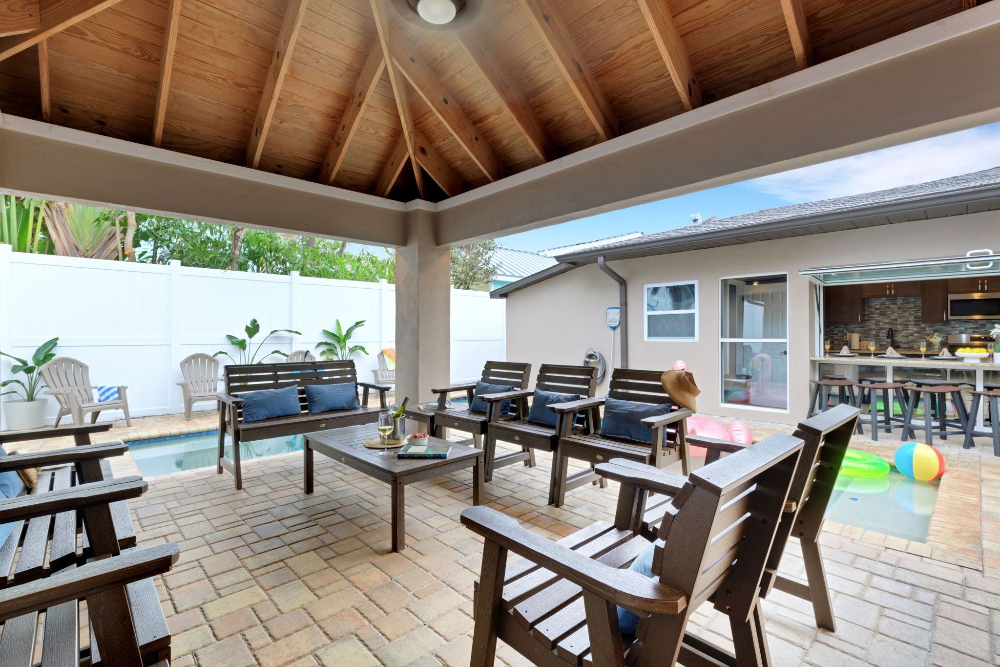 A covered patio with wooden furniture, a pool, and a kitchen area.