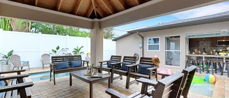 A covered patio with wooden furniture, a pool, and a kitchen area.