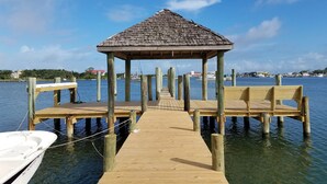 Un muelle de madera que lleva a un pabellón con techo de paja, rodeado de agua tranquila y cielo despejado.