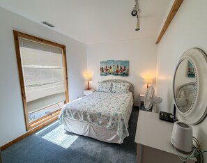 Ein Schlafzimmer mit Bett, Nachttisch, Lampe, Spiegel, Deckenventilator und einem Fenster mit Jalousien.