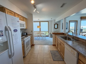 A modern kitchen with a refrigerator, microwave, stove, and sink. There is a dining area with chairs and a table, and a view of the outside through large windows. - Pirate's Quay 3 South:  Incredible harbor views and sunsets. Docking. (Ocracoke)