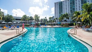 A swimming pool with lounge chairs and umbrellas, surrounded by palm trees and a tall building in the background. - Spacious condo w/ open layout across from the beach | WIFI + Parking (Sunny Isles Beach.)
