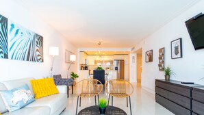 A modern living room with a white sofa, two rattan chairs, a glass coffee table, and a dark wooden sideboard. - Spacious condo w/ open layout across from the beach | WIFI + Parking (Sunny Isles Beach.)