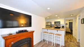 A modern kitchen with white cabinets, a microwave, and a built-in oven. There is a fireplace with a TV mounted above it, and a bar area with stools.