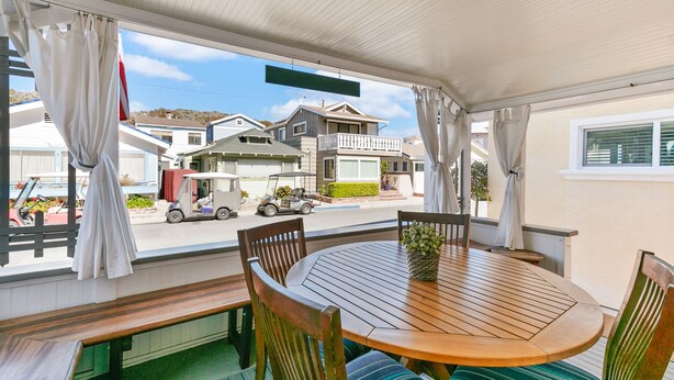 A covered patio with a wooden table and chairs, a bench, and a view of the street and houses.