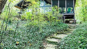Une maison moderne à la façade en verre, entourée d’une végétation luxuriante et dotée d’un escalier en pierre menant à l’entrée.