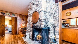Une cheminée en pierre avec un feu de bois, un plancher en bois et un coin cuisine visible à travers une porte ouverte.