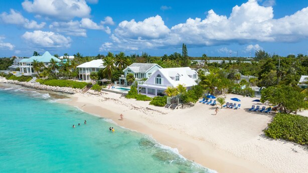 A beachfront property with a swimming pool, beach chairs, and a large house with a white roof.