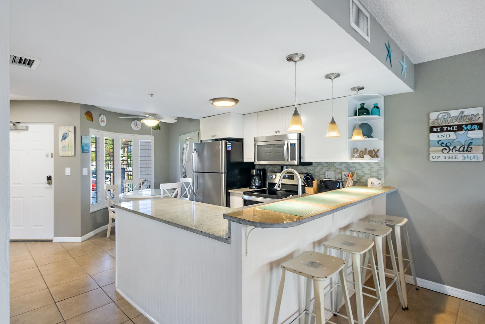 Een moderne keuken met een centraal eiland, barkrukken, roestvrijstalen apparatuur, hangende lampen en decoratieve wandkunst.