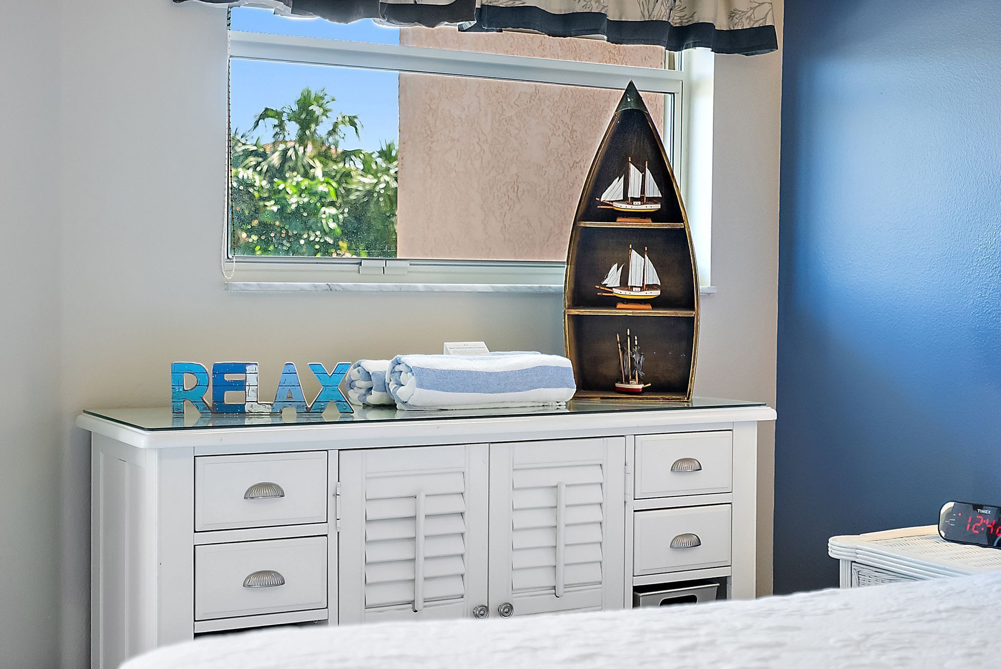 A bedroom with a white dresser, a window with a view of greenery, a shelf with a model ship, and a 'RELAX' sign.