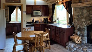 L’intérieur d’un chalet rustique comprend une table à manger en bois, un coin cuisine, une cheminée en pierre et un tableau encadré accroché au mur.