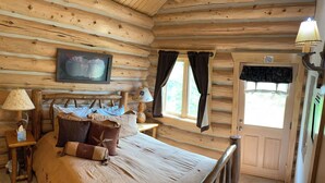 Une chambre dans un chalet en rondins, avec un lit, des oreillers, une télévision et une fenêtre avec des rideaux.