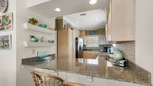 A kitchen with granite countertops, wooden cabinets, and a central island with two wicker chairs. - "Jen's Creole Cottage" Amazing Gulf Front Property! (Navarre Beach)