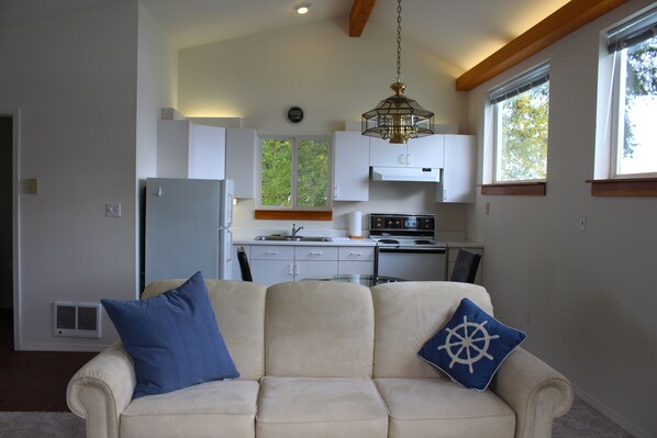A living room with a beige sofa, a blue pillow, and a ship wheel pillow. A kitchen is visible in the background with white cabinets and a stove.