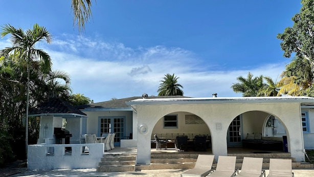 Zona de piscina con sillones, un área de estar cubierta y un edificio blanco con arcos.